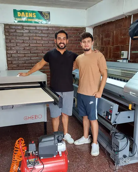 Nuestro taller en 2023: dos personas junto a la plancha y el plotter, con el cartel de Daens en la pared